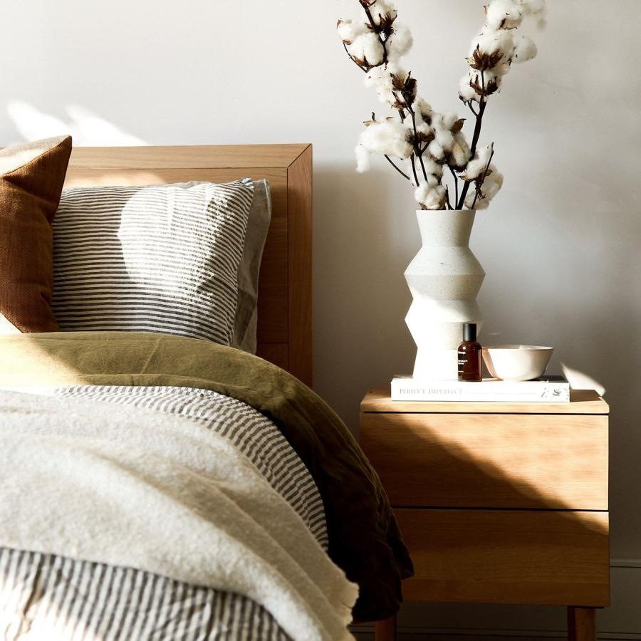 swath of sunlight shines across a well made bed and bedside table with a vase of cotton plants
